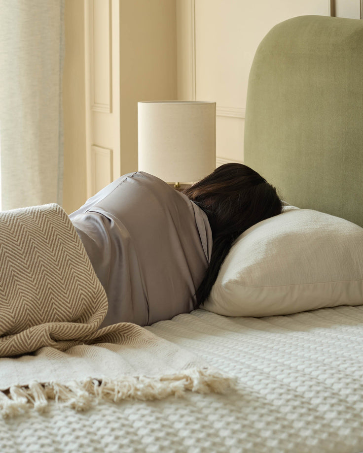 Close-up of a contemporary upholstered bed with a detailed headboard featuring vertical stitching and a quilted fabric pattern, in a soft neutral colour palette, set against a warm, ambient bedroom interior with a wooden door and a table lamp in the background.