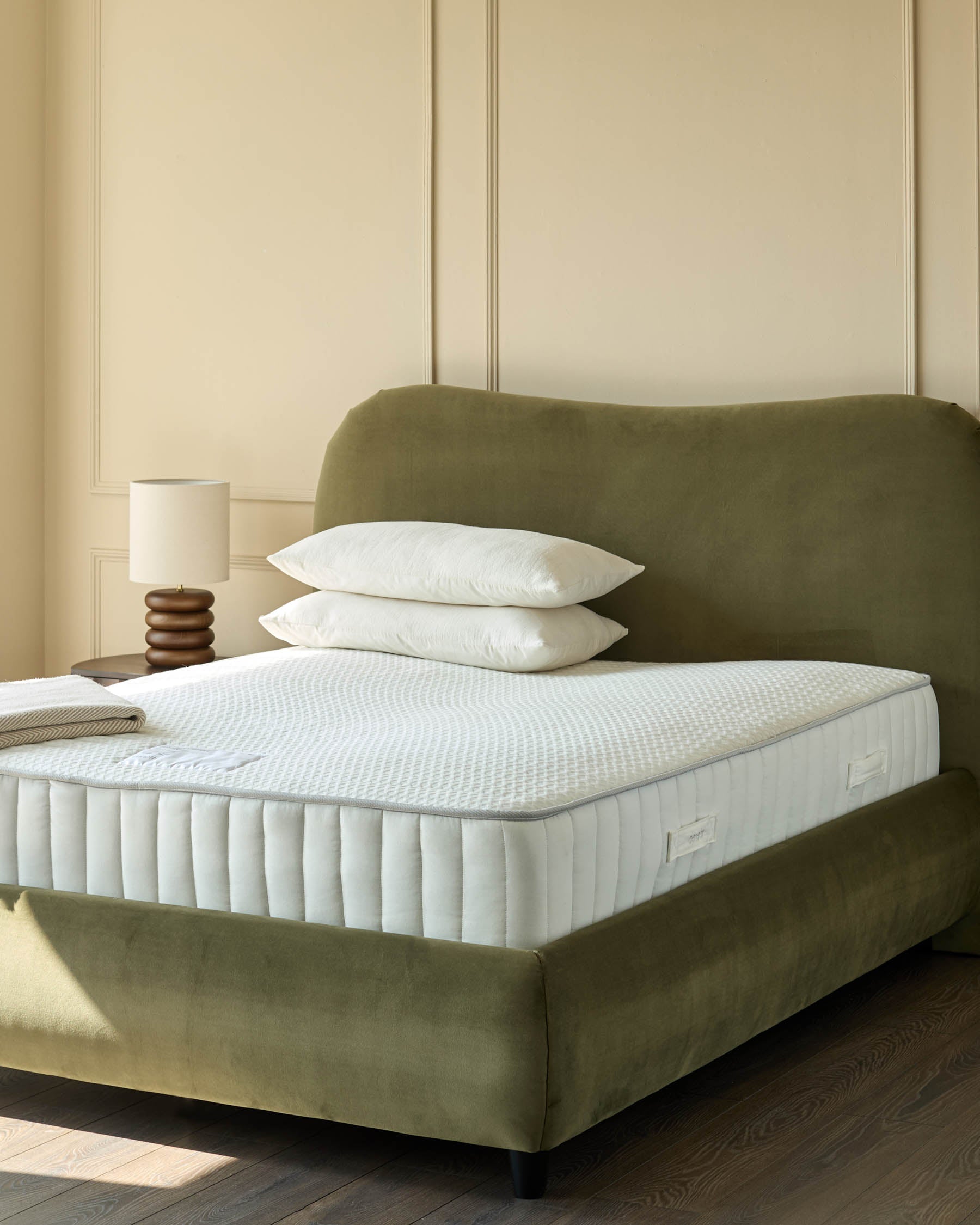 An olive green velvet bed with a white mattress and pillows sits in a room with a beige wall and wood floors.