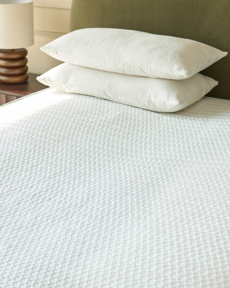 A bedroom scene with pillows on a white waffle-textured bed and a wooden bedside lamp.