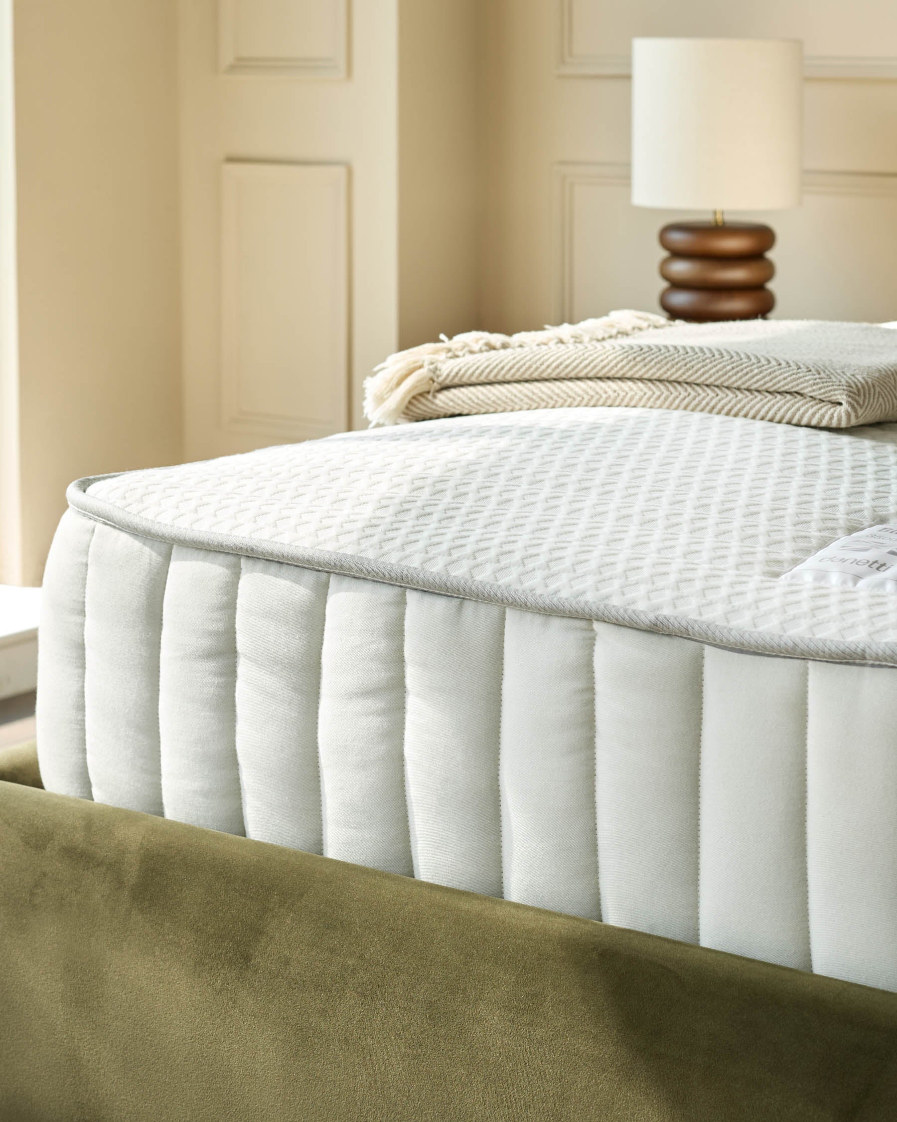 Close-up of a mattress with white quilted cover and olive green base with a lamp and folded blanket in the background.