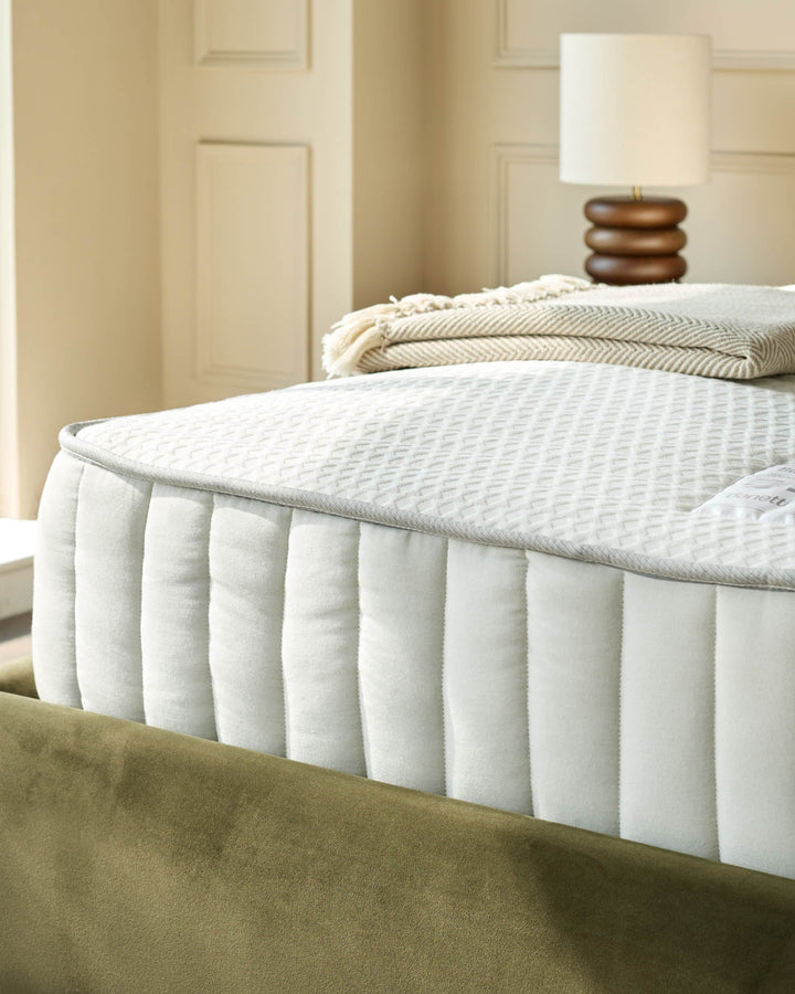 Close-up of a mattress with a white cover, textured pattern, and green velvet base. Lamp and throw blanket in the background.