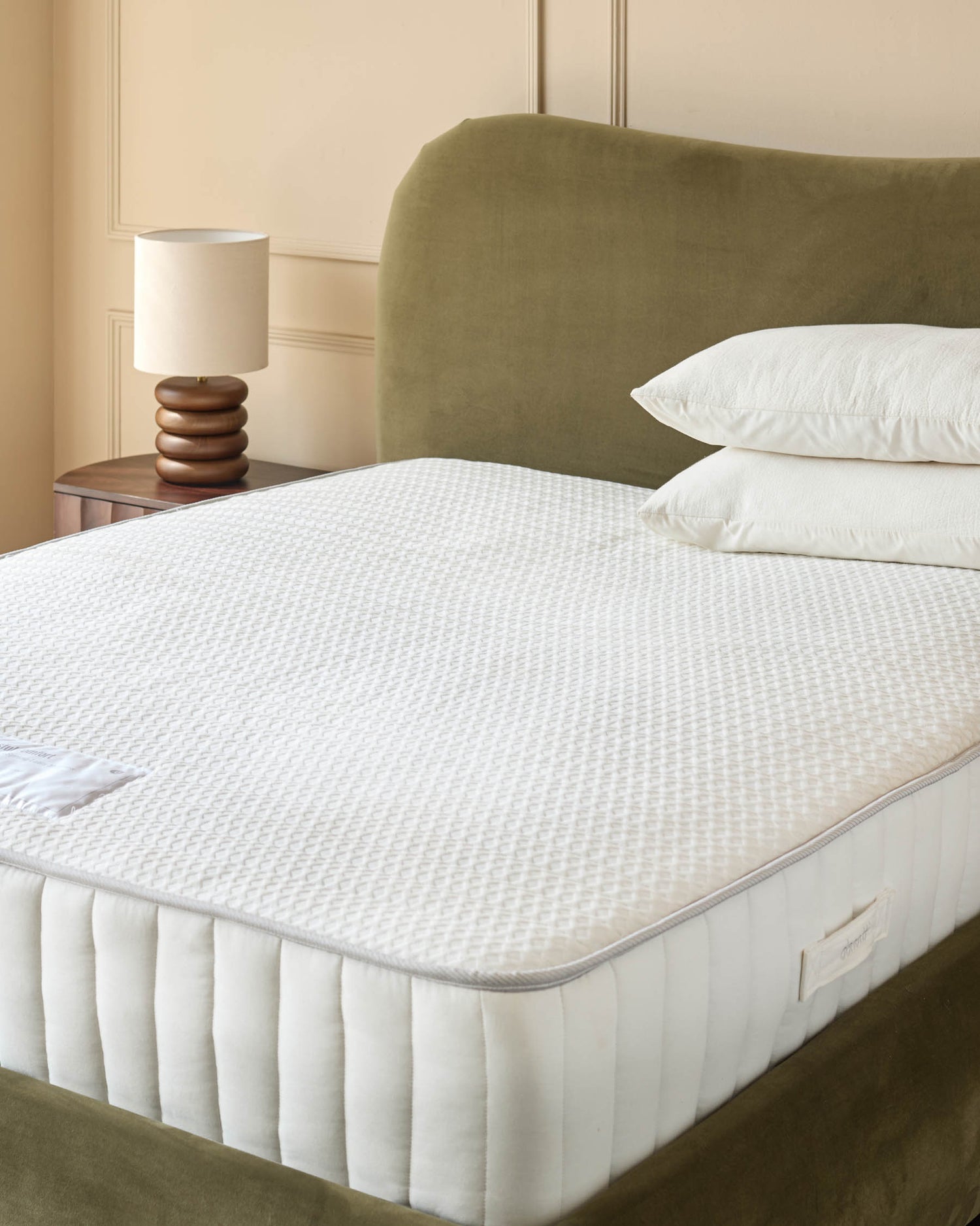 A bed with a white mattress, two white pillows, and an olive-green headboard. A wooden bedside table and lamp are visible.