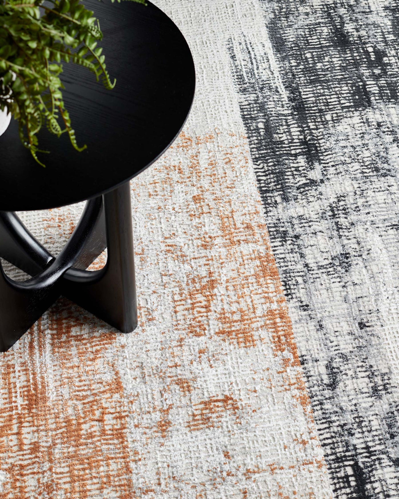 A modern circular black side table with three intersecting legs, partially displaying a fresh green fern on top. The table rests on a contemporary area rug with an abstract design featuring white, grey, and terracotta hues.