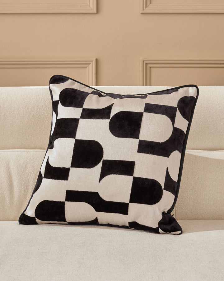 Black and cream geometric patterned cushion with piping detail, displayed on a beige sofa against a neutral wall.