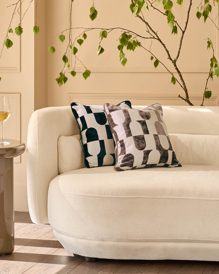 Bridget cushion in chocolate brown and cream geometric pattern, displayed on a cream curved sofa with decorative styling.