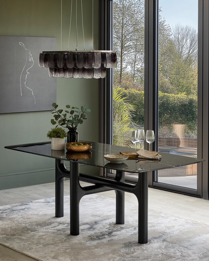 A modern dining table with a sleek black glass top and statement dark wood legs, complemented by chic decor and natural light.
