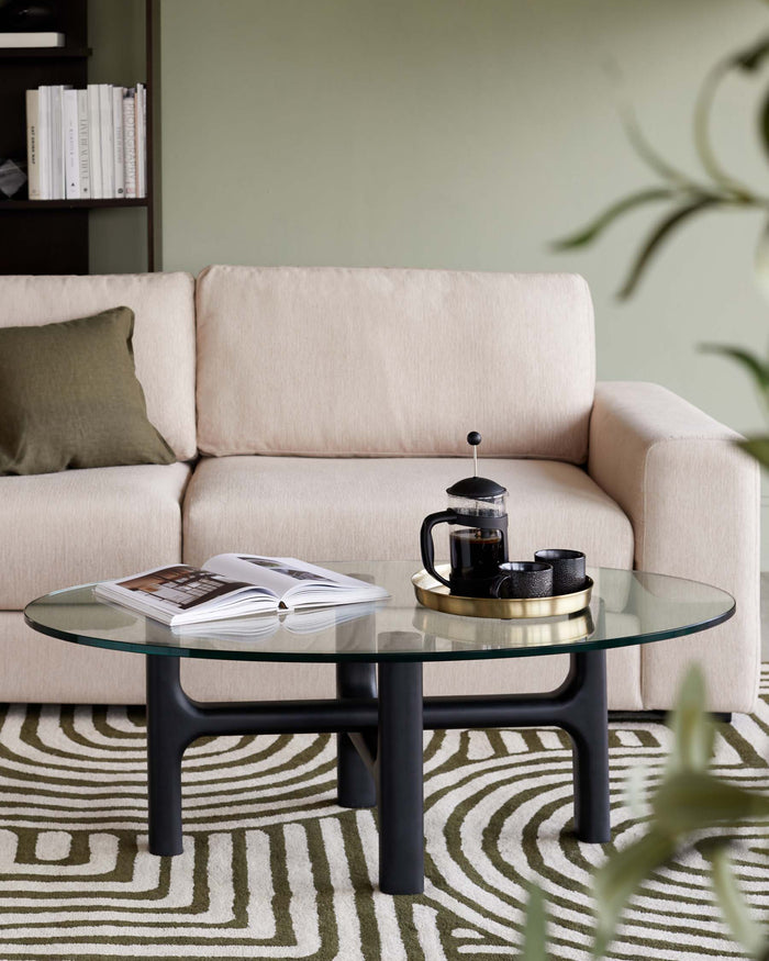 A contemporary beige fabric three-seater sofa with plush cushions, paired with a modern oval glass-top coffee table featuring a matte black base. The setting is complemented by a geometric-patterned area rug in neutral tones.