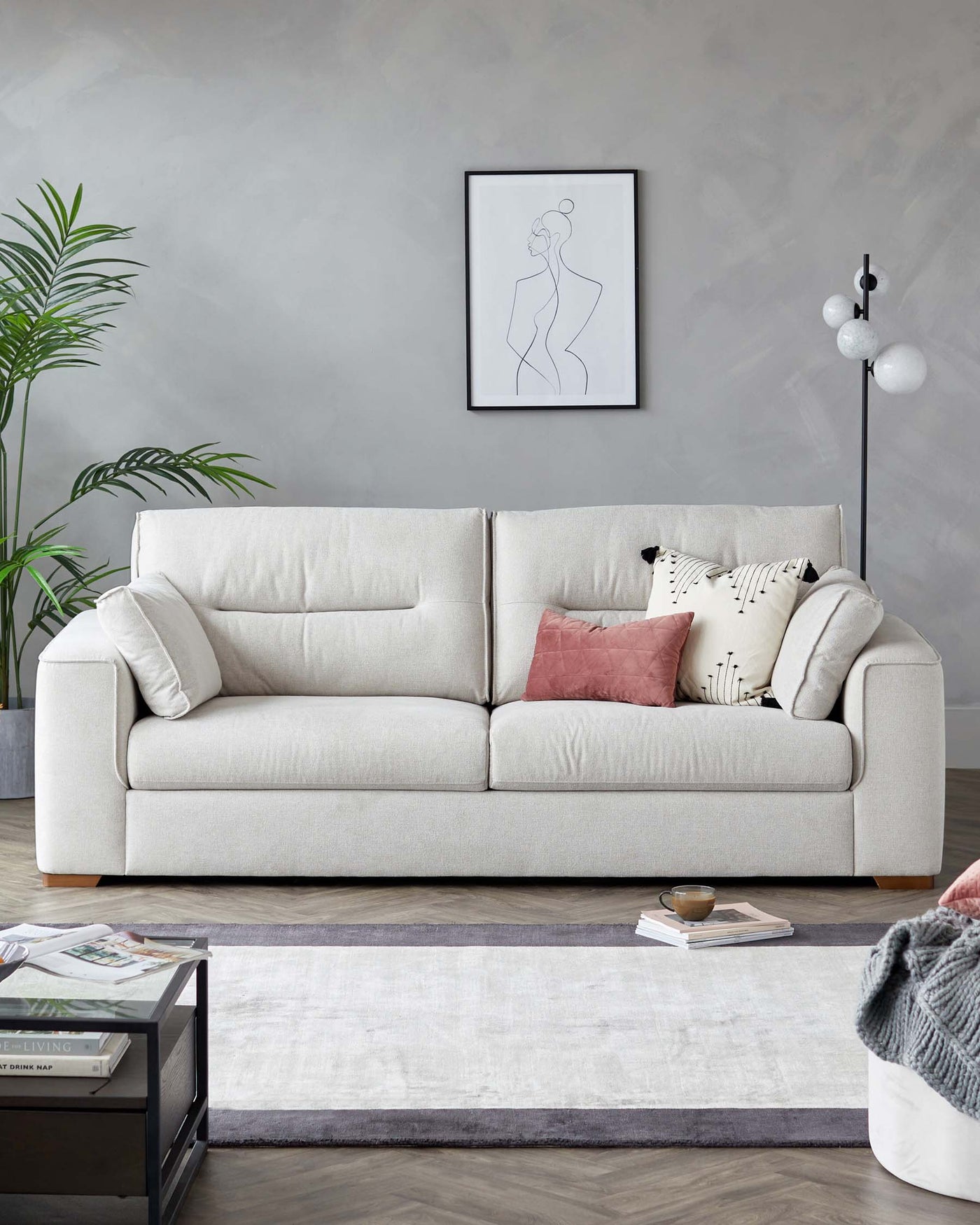 A contemporary light grey fabric three-seater sofa with plush cushions, flanked by patterned and solid-coloured decorative throw pillows. In front of the sofa lies a neutral colour area rug, and to the side, a modern black floor lamp with spherical white shades.