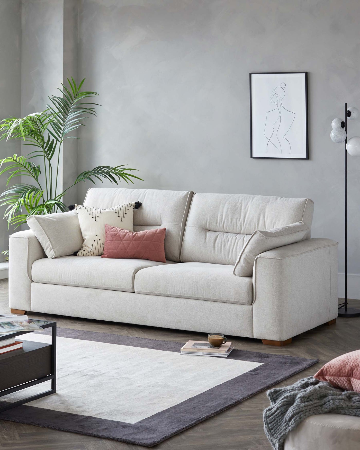 A contemporary light beige fabric three-seater sofa with cushions in various shades of pink and beige. In front of the sofa is a modern black frame coffee table with a glass top, over a two-tone grey and white area rug. The setting is complemented by a tall floor lamp and a framed line art piece on the wall.