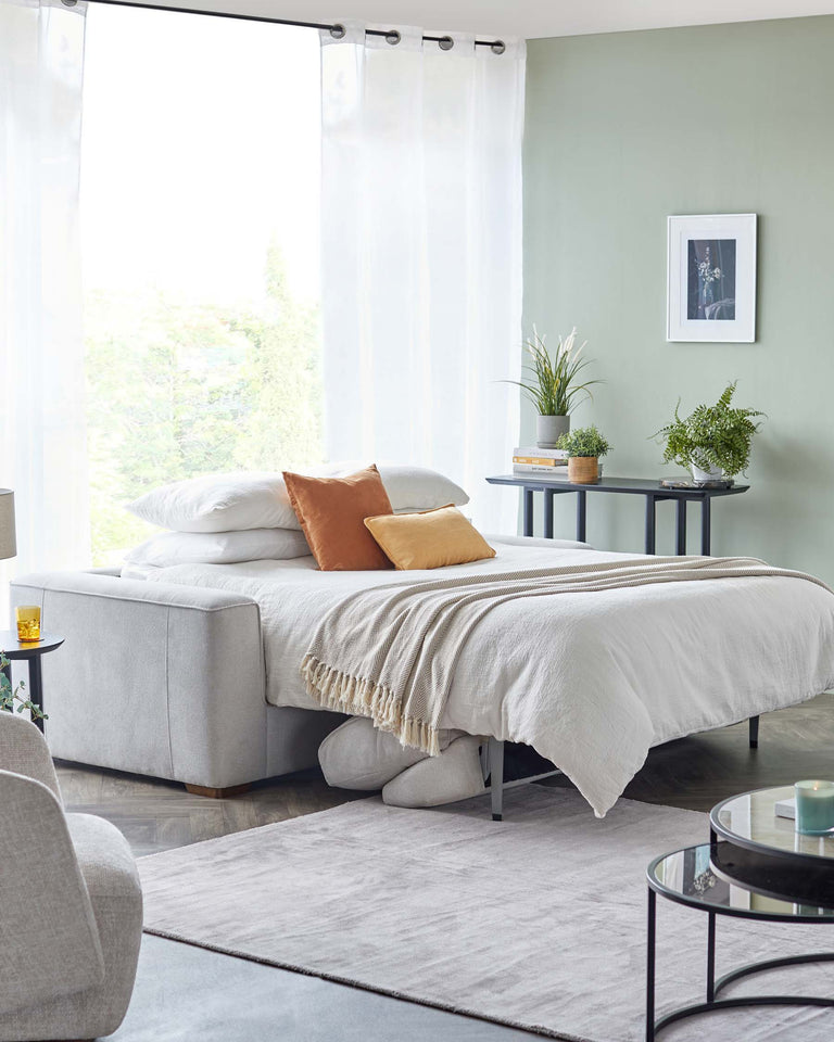 Contemporary styled bedroom featuring a light grey upholstered bed with plush white bedding and a variety of pillows, including a beige throw pillow with a fringe detail. A round, black metal framed coffee table with a glass top sits on a soft grey area rug, accompanied by a minimalist grey armchair. A sleek, round side table with two levels holds decorative plants and books, creating a refreshing ambiance. Transparent white curtains allow natural light to enhance the serene colour palette of the room.