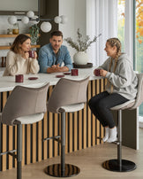 Bronwyn gas lift bar stool in natural and mocha velvet, shown in kitchen setting with three people seated at counter.