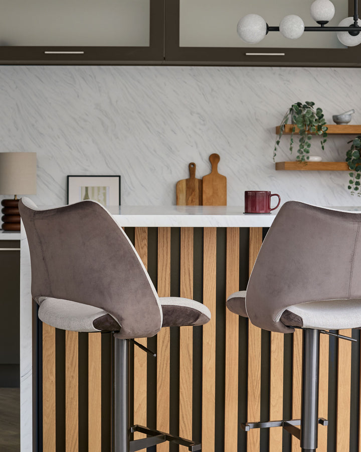 Bronwyn gas lift bar stools in mocha velvet and natural fabric, shown at kitchen counter with wood and black striped base detail.