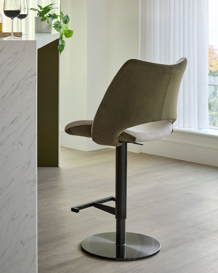 Bronwyn gas lift bar stool in olive green velvet and natural fabric with black metal base, shown in modern interior setting.