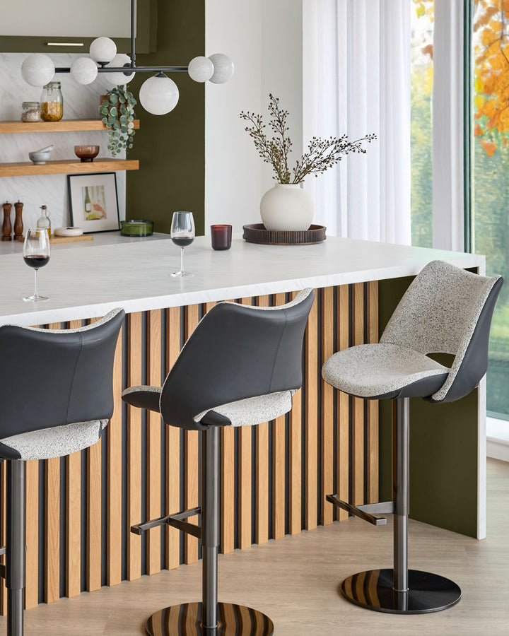 Bronwyn gas lift bar stool in grey fabric and black faux leather, shown at white kitchen island with striped wood paneling.