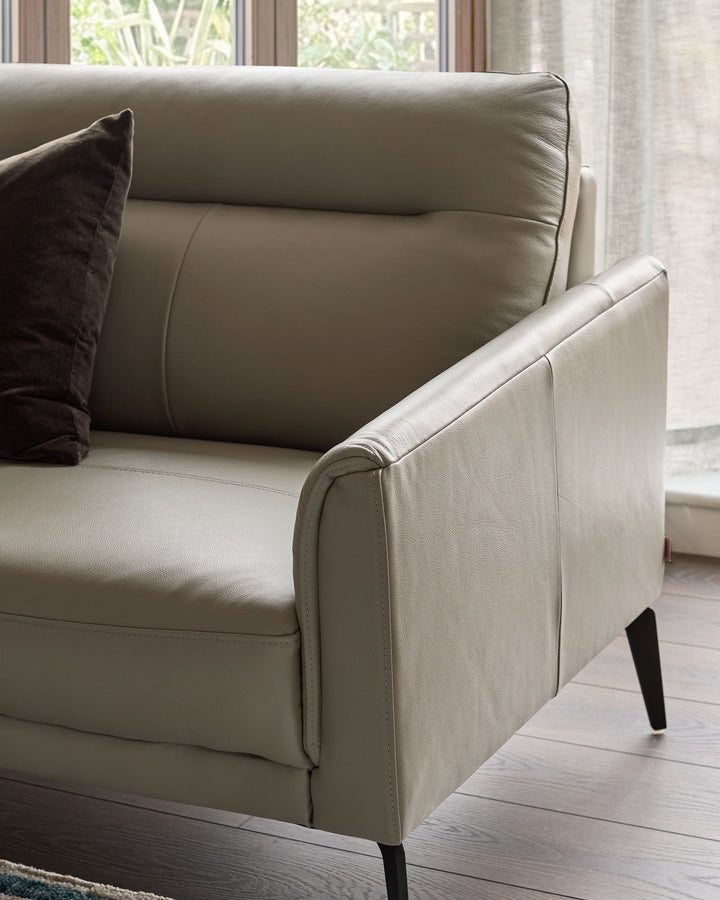 Light grey leather left-hand corner chaise sofa with curved armrest, cylindrical back cushion, and black tapered legs, positioned near windows.