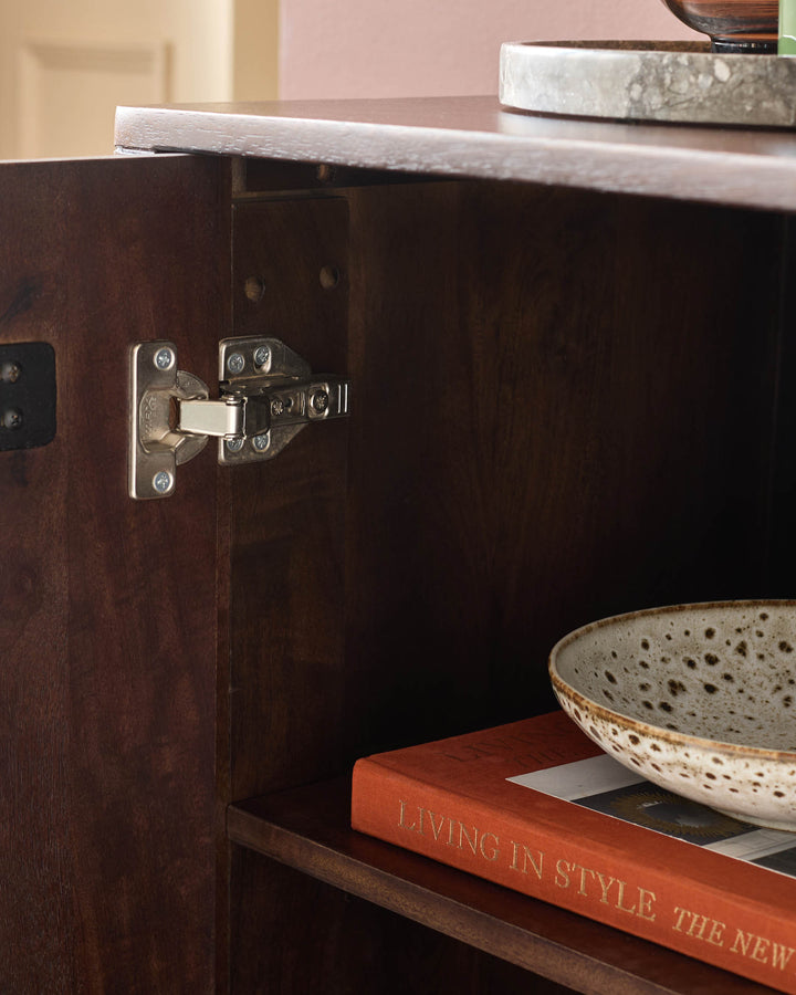 Close-up of a cabinet with door ajar showing a hinge. On an interior shelf, there is a bowl and a book.
