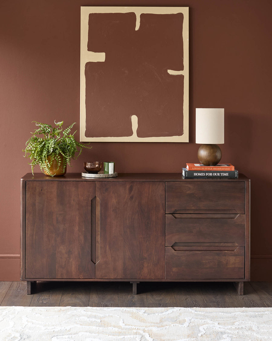 A stylish dark wood sideboard with drawers, a lamp, books, and a green plant atop against a brown wall and floor.