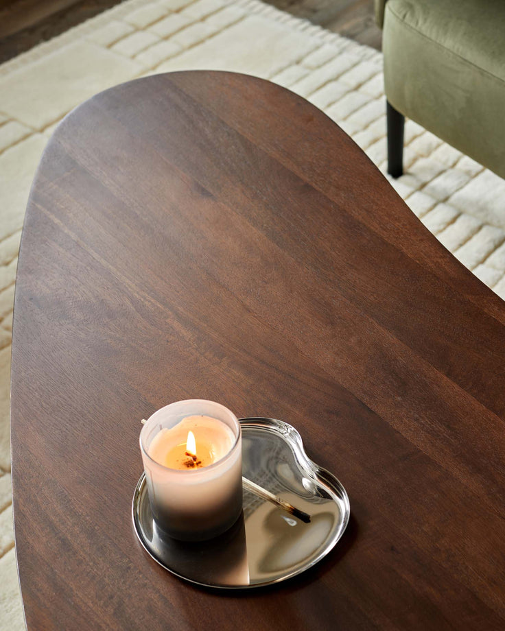 A wooden coffee table with a lit candle sat on a shiny silver coaster. The table is on a neutral rug with an olive green chair.