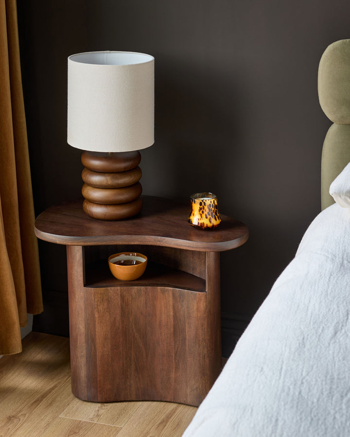 Dark mango wood side table with curved top, open shelf, and rounded base, styled in a bedroom setting.