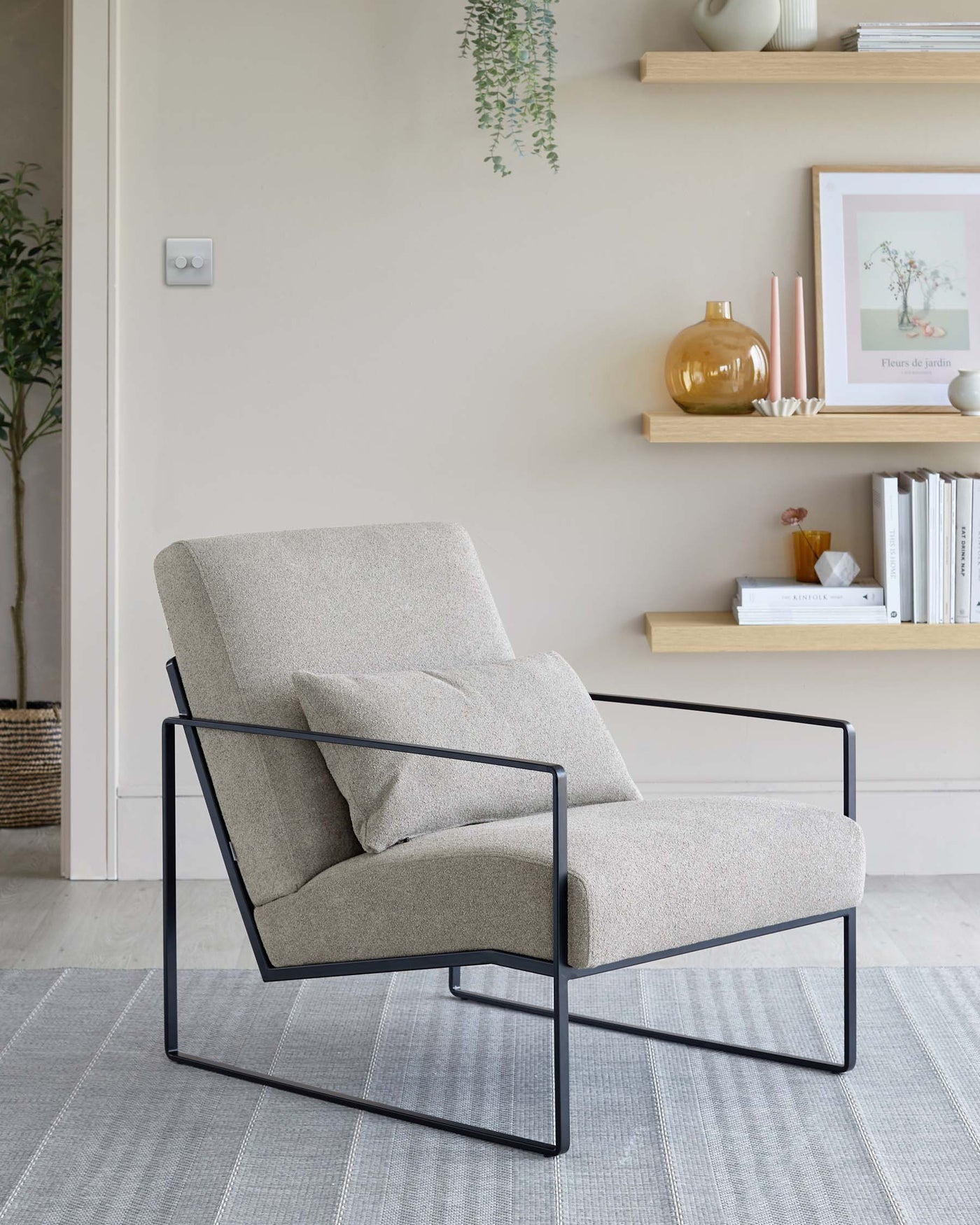 A contemporary light beige fabric upholstered armchair with a minimalist black metal frame, featuring a comfortable backrest and a cushioned seat with a pillow, set against a neutral-toned room with decorative shelving on the wall.