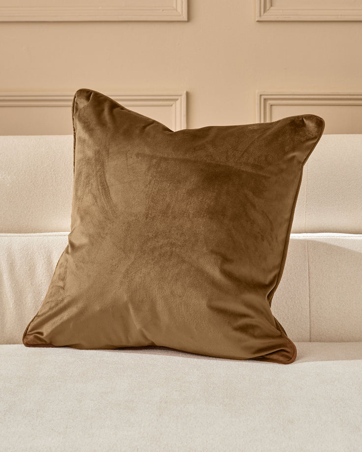 Micah medium cushion in chocolate brown velvet displayed on a cream upholstered sofa against a beige paneled wall.