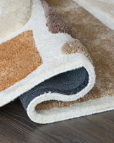 A plush, multi-toned rug with neutral hues, partly rolled to reveal its textured backing on a dark wooden floor.