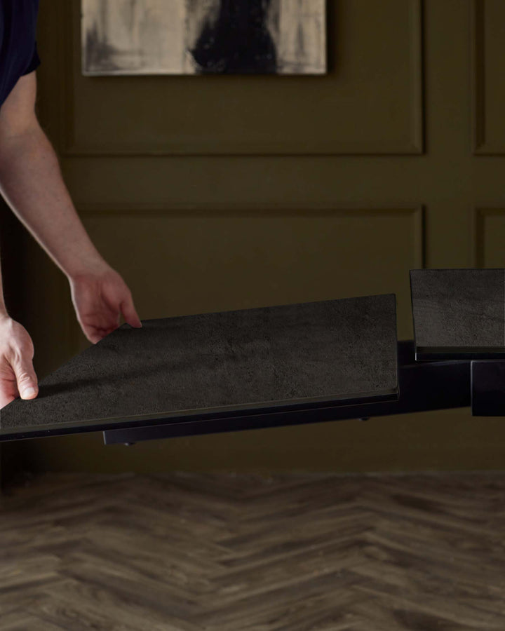 A modern, minimalistic dining table with a dark textured top and sleek edges, being adjusted with an additional panel.