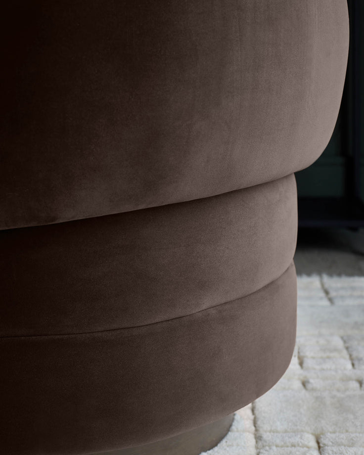 A close-up of a chocolate brown velvet pouffe, showing the plush texture and curved form of the furniture.
