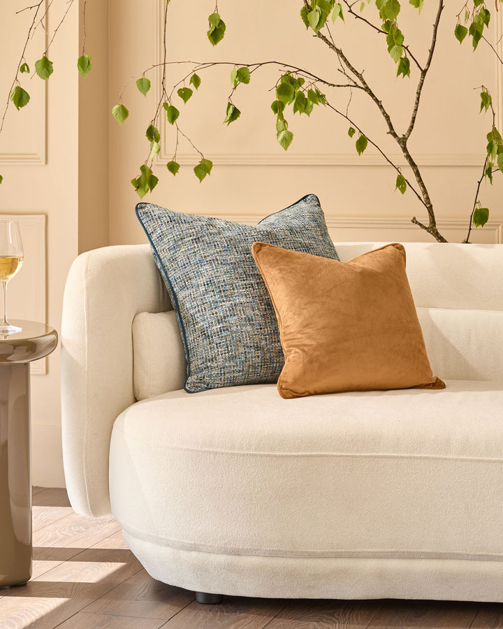 Caramel leather cushion displayed on a cream curved sofa with decorative branches above.