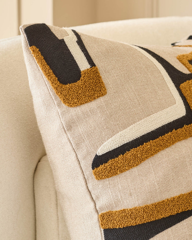 Camille cushion in natural with geometric pattern in mustard, black and cream, displayed on beige sofa.