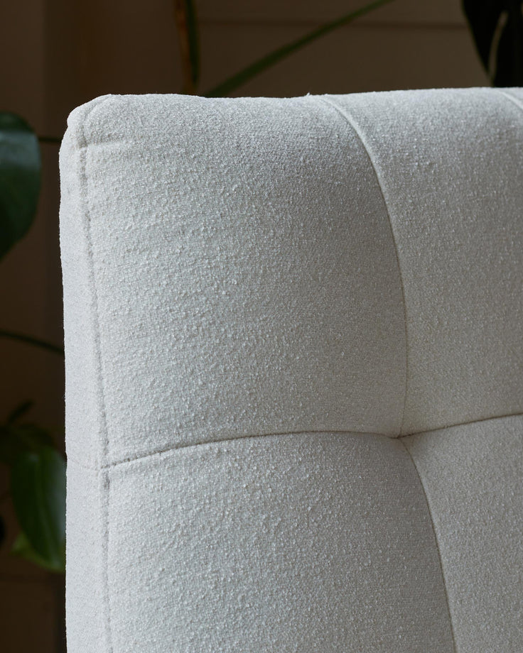 Close up of a textured, off-white, upholstered chair back, with button detail, against a blurred, leafy background.
