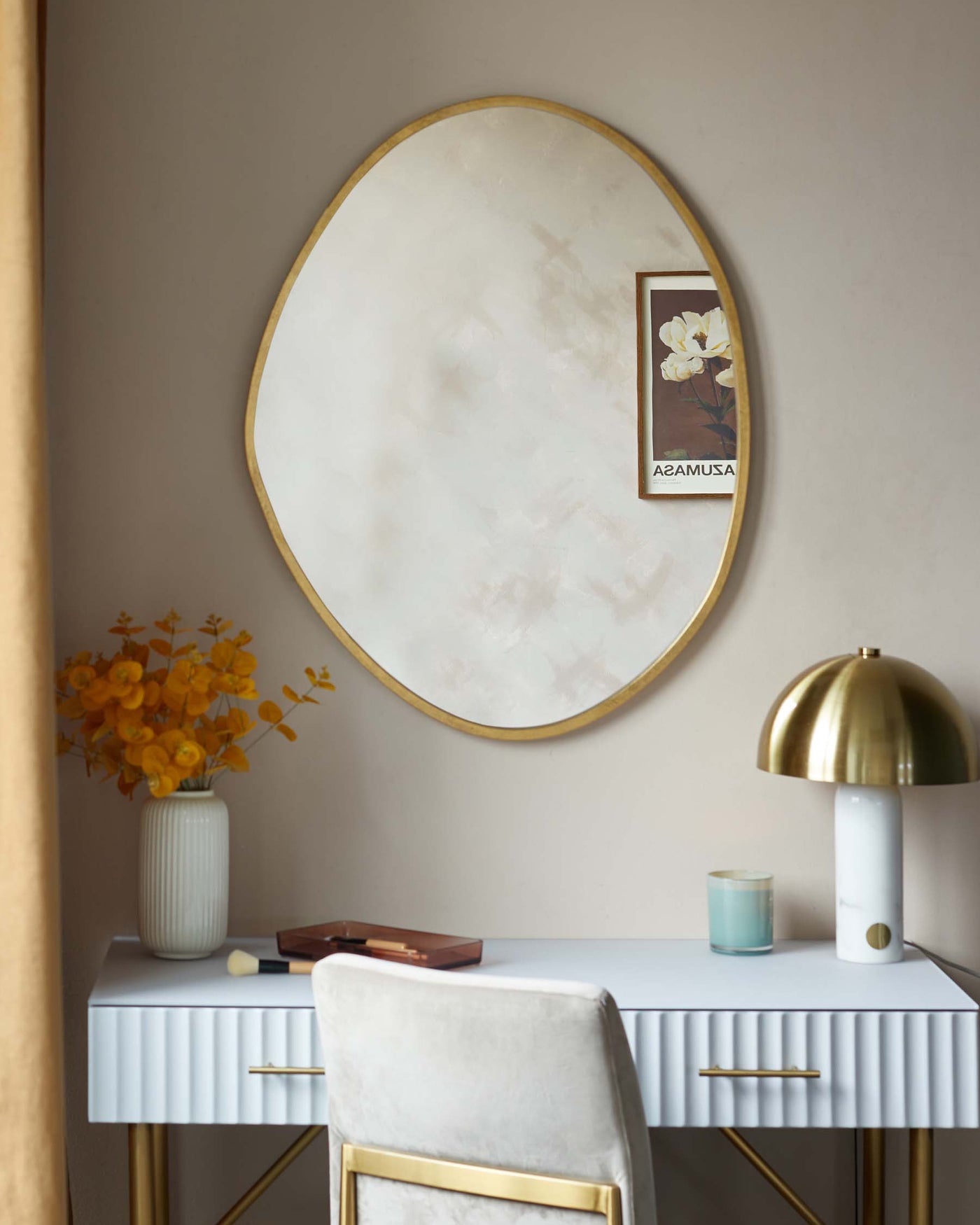 Elegant modern vanity desk with a white and pleated texture finish and gold handles, accompanied by a matching stool with a white cushion and gold frame. The furniture is complemented by a large, oval-shaped mirror with a subtle gold frame on the wall.