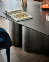 Carlo 8-seater dining table in dark grey ceramic with cylindrical base, styled with magazines and candles.