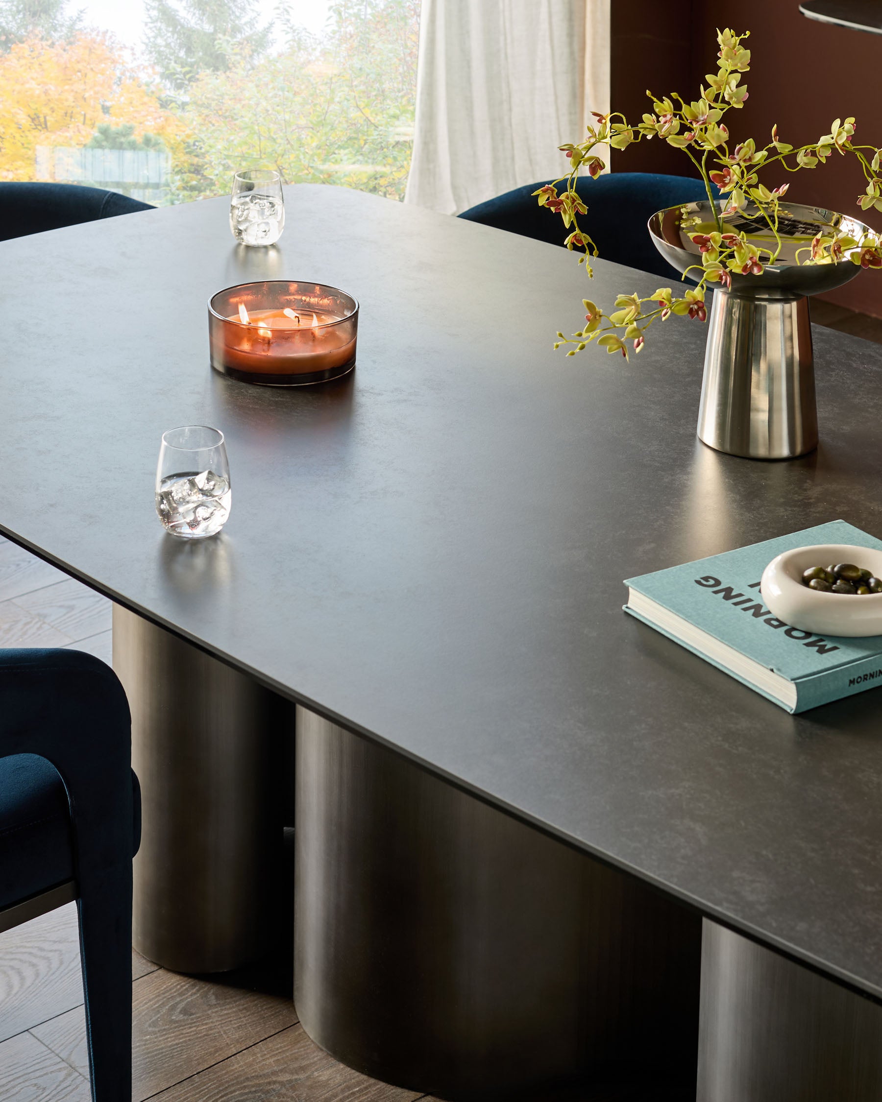 Carlo 8-seater dining table in dark grey ceramic with curved metal base, styled with chairs, candle, and flowers.