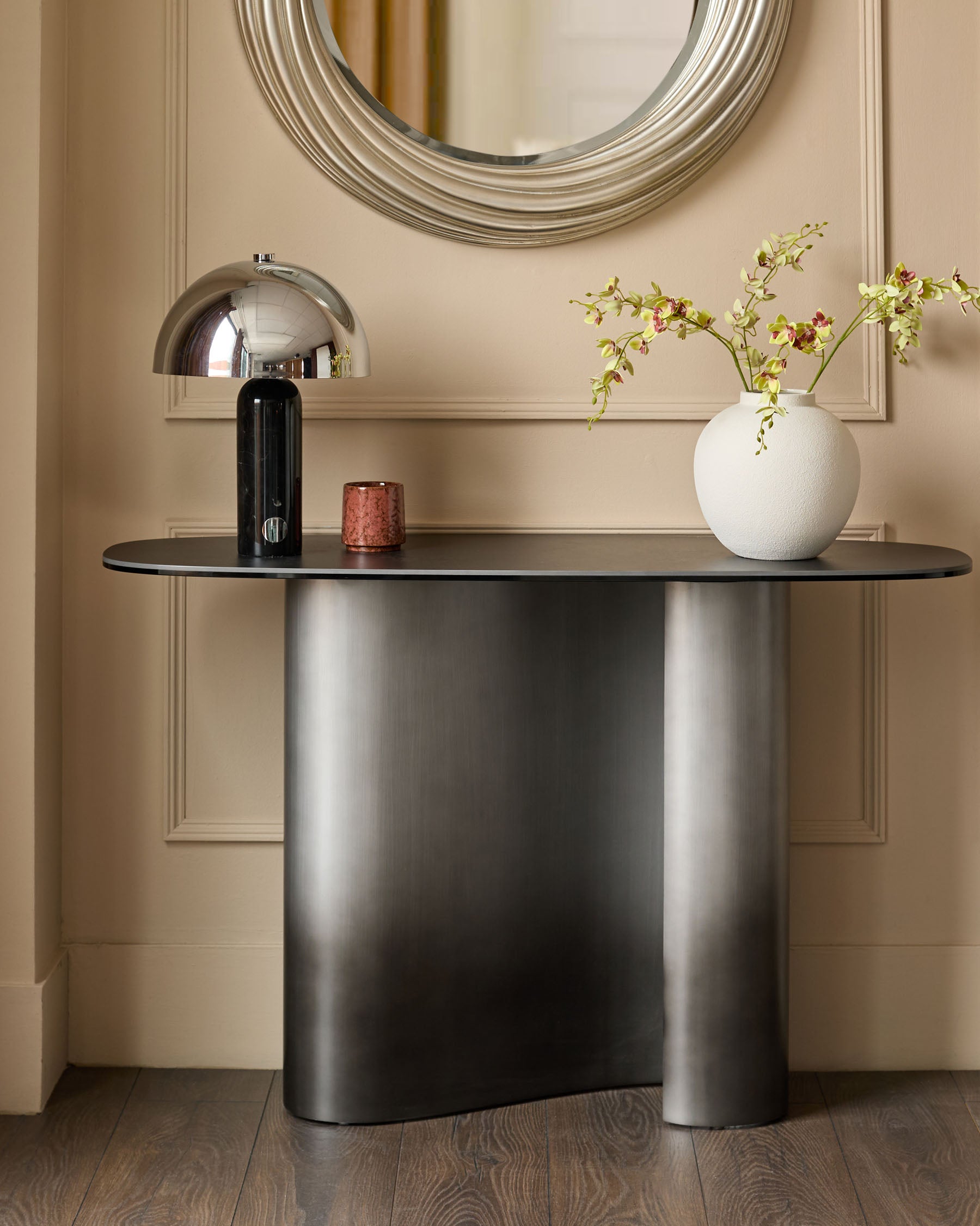 Carlo console table in dark grey ceramic with curved base, styled with chrome lamp and white vase in contemporary entryway setting.