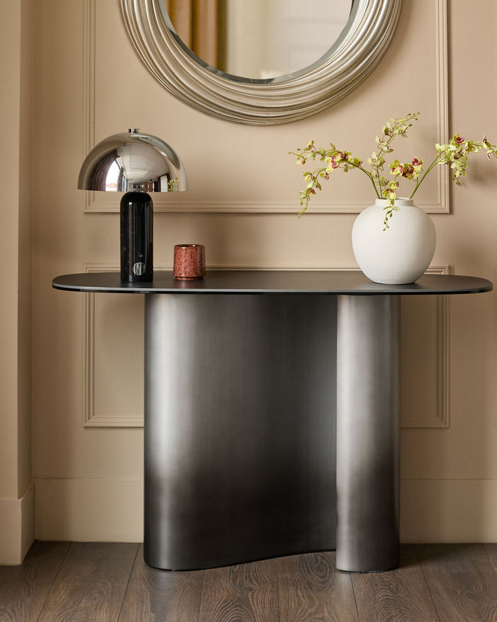 Carlo console table in dark grey ceramic with curved base, styled with chrome lamp and white vase in contemporary entryway setting.