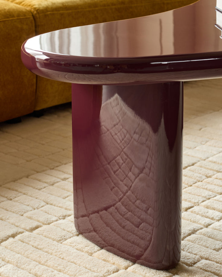 Gloss plum coffee table with curved top and textured pedestal base, paired with mustard chair on cream rug.