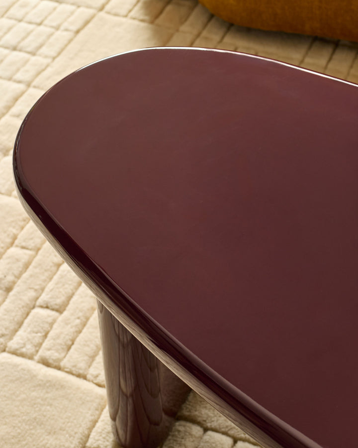 Carmela coffee table in gloss plum, shown from above at an angle to highlight its curved edges and reflective finish on a cream brick floor.