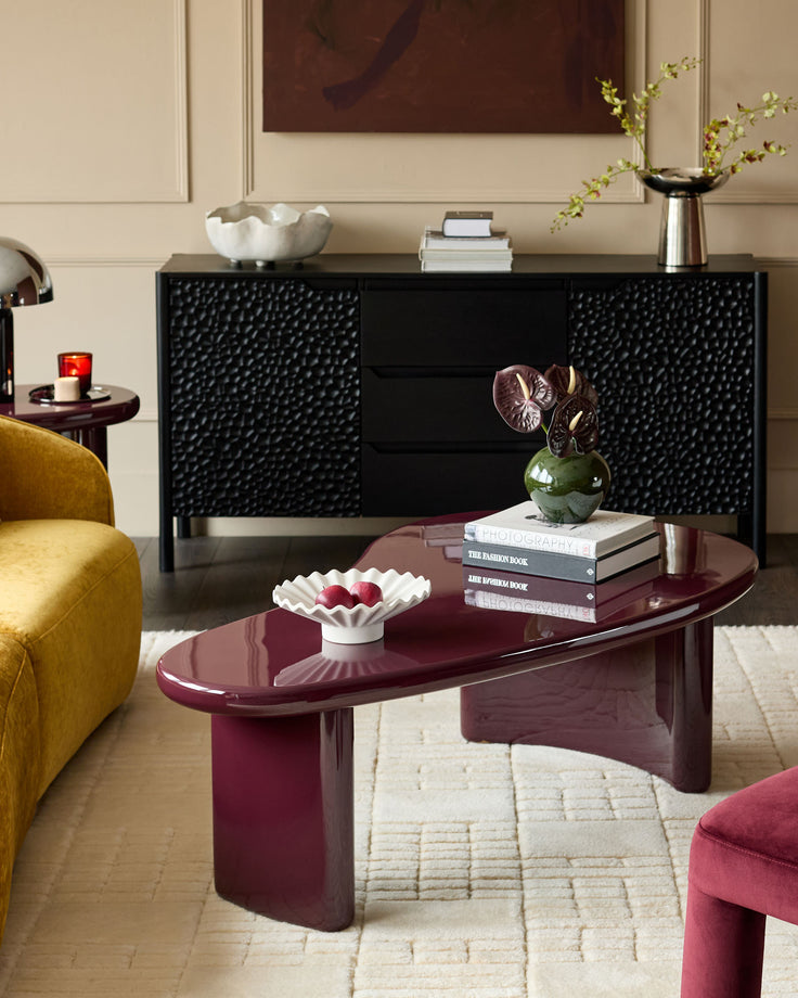 Gloss plum coffee table with curved oval top and sculptural pedestal base, styled in modern living room setting.