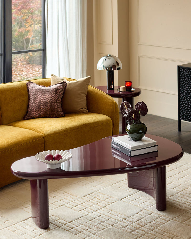 Carmela coffee table in gloss plum finish with organic rounded oval shape, displayed in a styled living room setting.