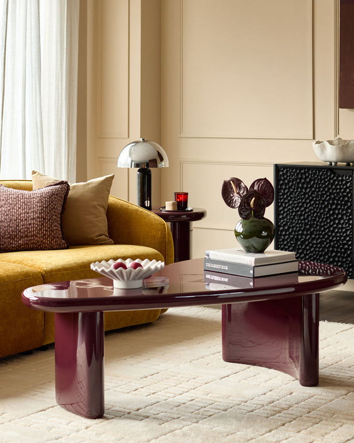 Carmela coffee table in gloss plum finish, oval shape with sculptural curved legs, displayed in a modern living room setting.