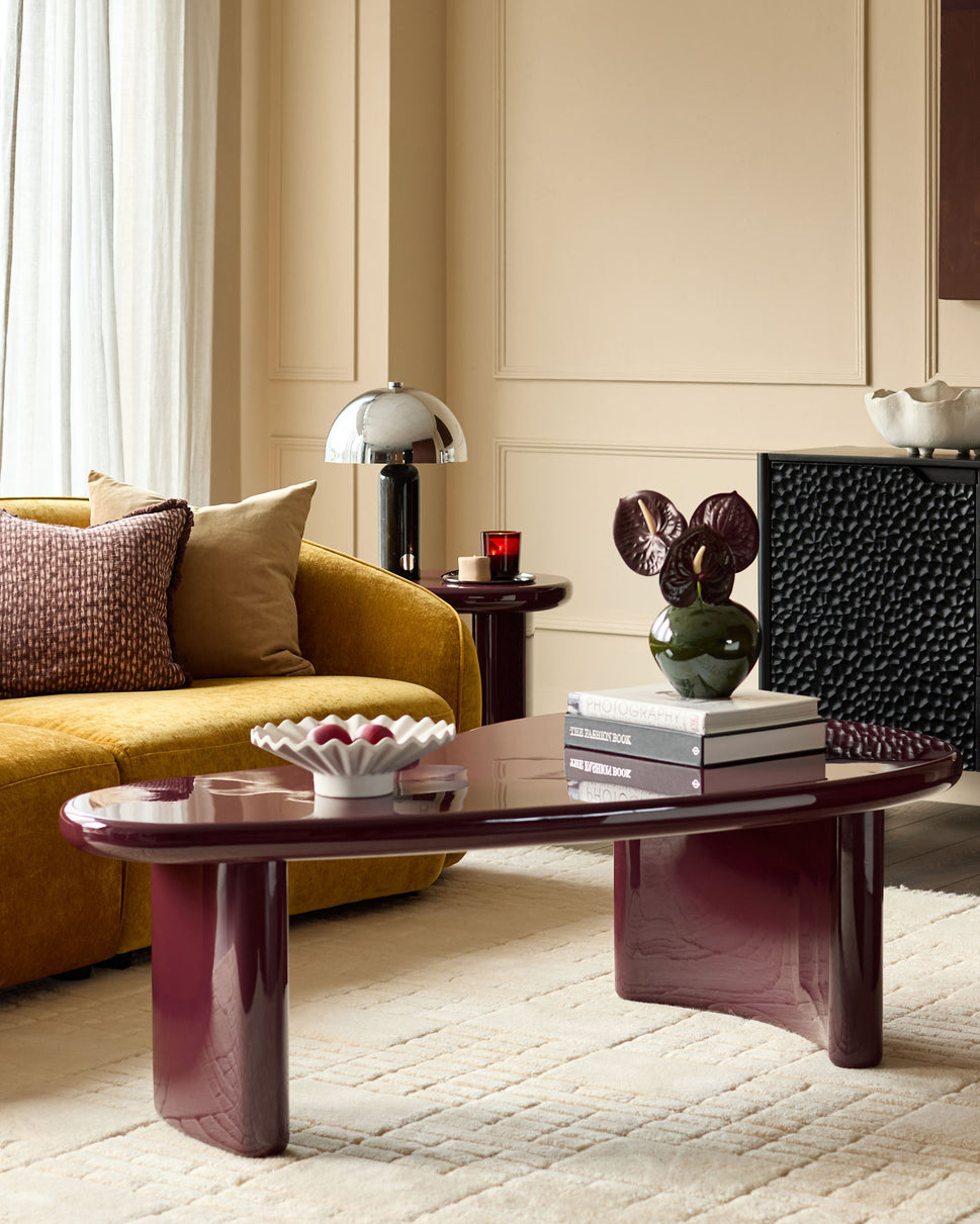 Carmela coffee table in gloss plum finish, oval shape with sculptural curved legs, displayed in a modern living room setting.