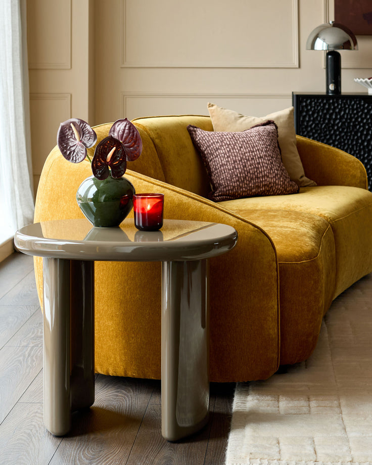 Carmela side table in gloss olive green, shown as a curved, sculptural accent table next to a mustard yellow sofa in a modern living room.