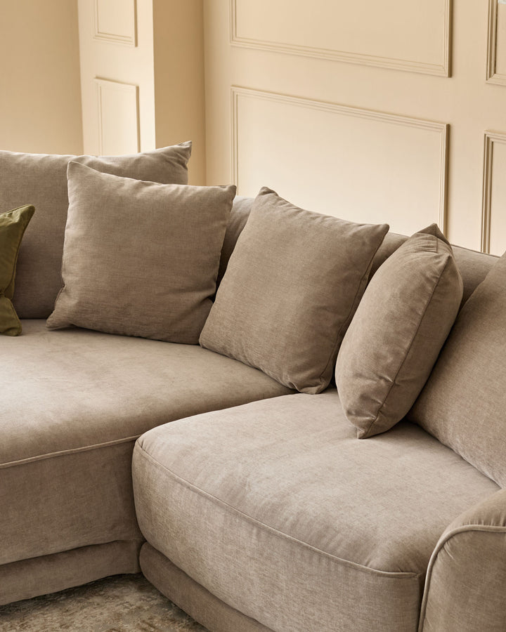 Caspian left-hand corner chaise sofa in mink fabric, angled view showing curved seating with scatter cushions against panelled wall.