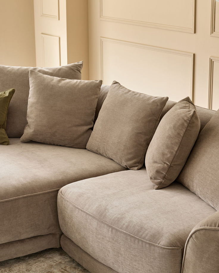 Caspian left-hand corner chaise sofa in mink fabric, angled view showing curved seating with scatter cushions against panelled wall.