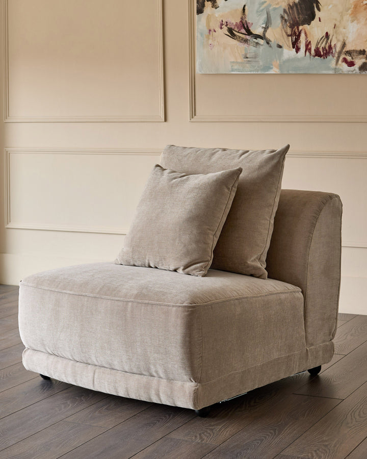 Mink fabric single module chair with two decorative pillows on dark wood flooring against paneled wall.