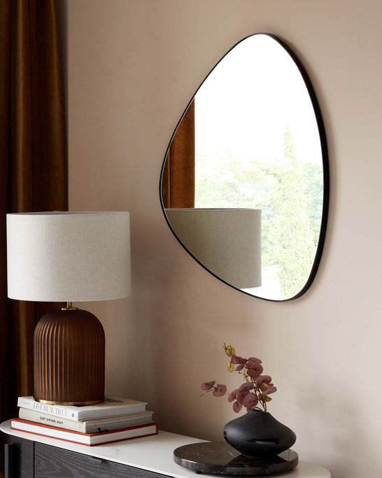 A modern, teardrop-shaped table lamp with a ribbed bronze base and a white drum shade, placed on a stack of decorative books on a sleek white console table. A simple, organic-shaped black vase with pink foliage complements the setup, under a large, ovular wall mirror with a thin black frame, reflecting a serene outdoor view.
