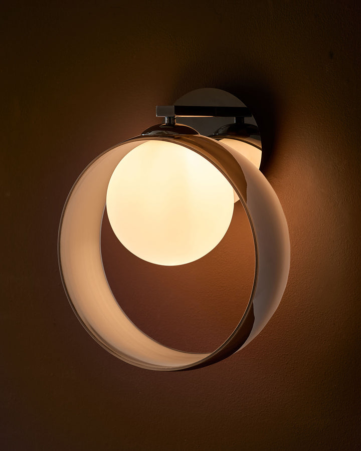 Modern circular wall sconce with frosted glass shade, chrome fixture, and warm ambient lighting against textured brown wall.