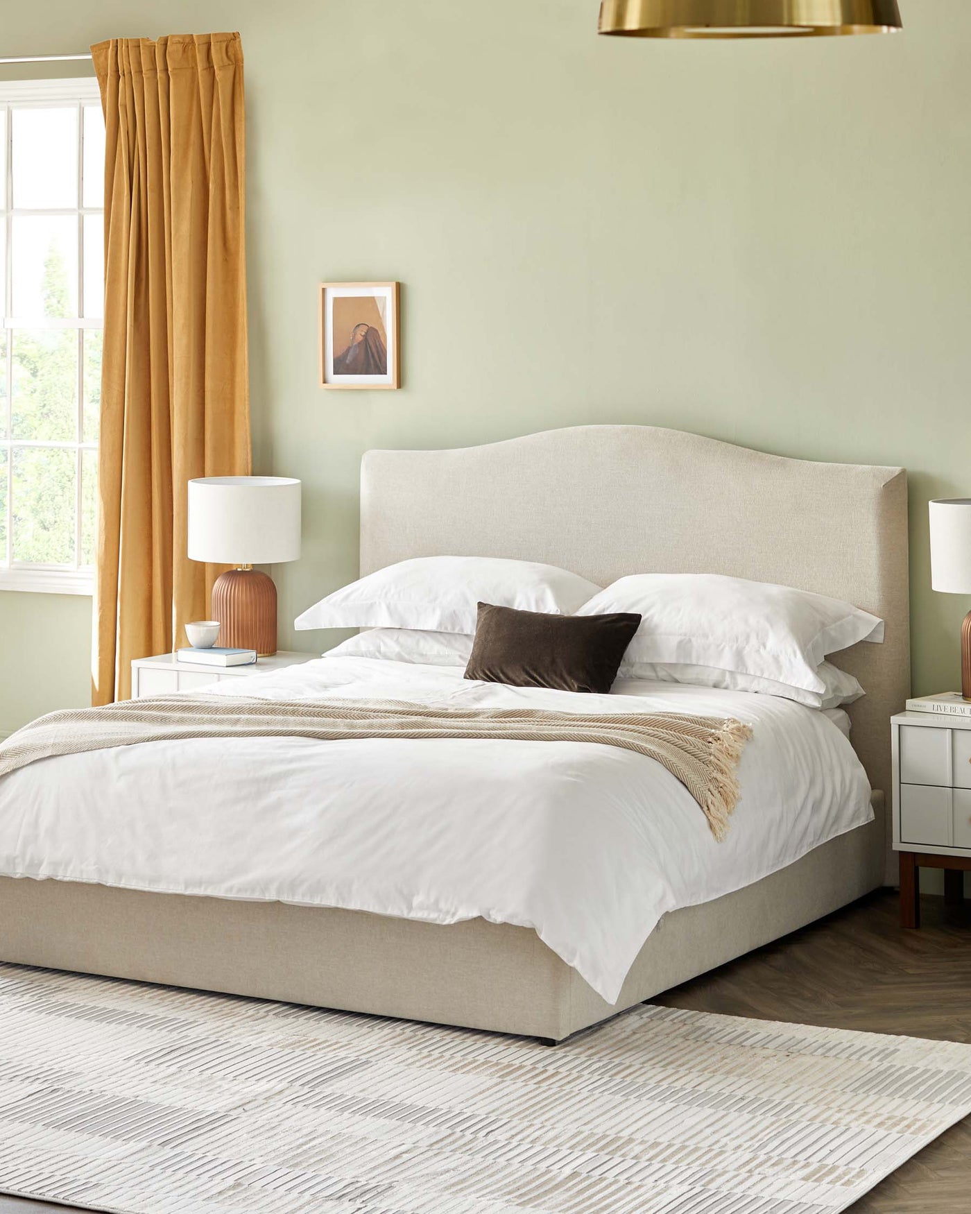 Beige upholstered bed frame with a curved headboard, white bedding, and accent pillows. Two white bedside tables with a wood finish top and storage drawers. A beige area rug with a white pattern.