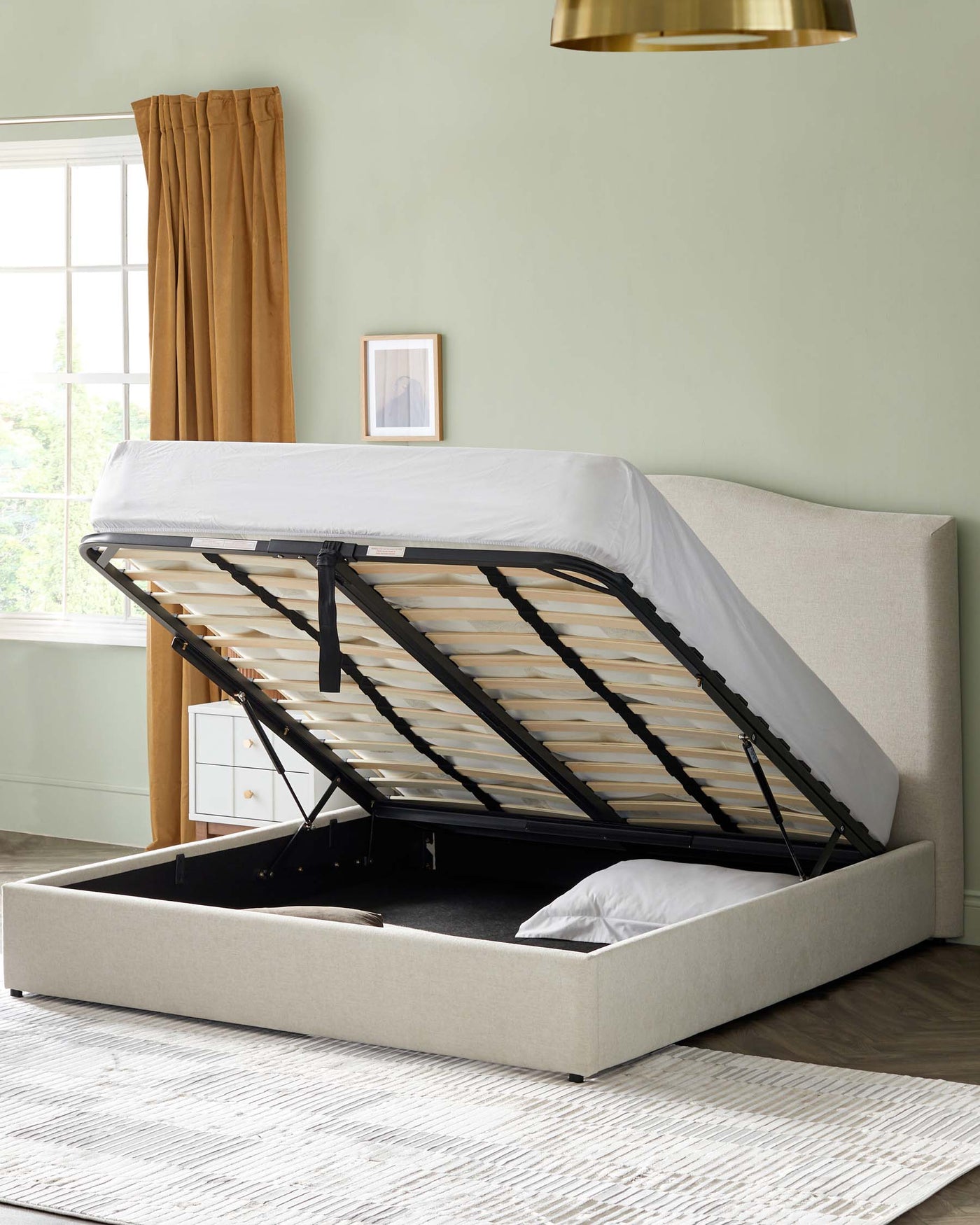 Beige upholstered storage bed with a gas-lift mechanism showcasing the under-bed storage area and a slatted bed base, partially covered with a white sheet, in a room with a green wall and wooden flooring.